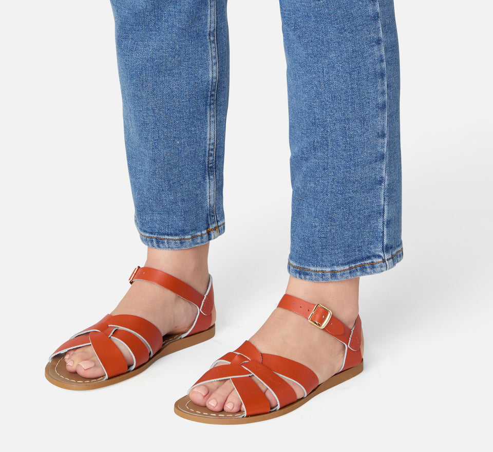 An original girls sandal by Salt Water Sandals in paprika with a single buckle fastening around the ankle. Open Toe and Sling-back with woven detail on the front. Lifestyle of standing view.