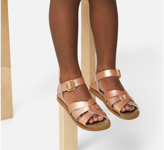 An original girls sandal by Salt Water Sandals in rose gold with single buckle fastening around the ankle. Open Toe and Sling-back with woven detail on the front. Lifestyle of girls legs dangling from a chair view.