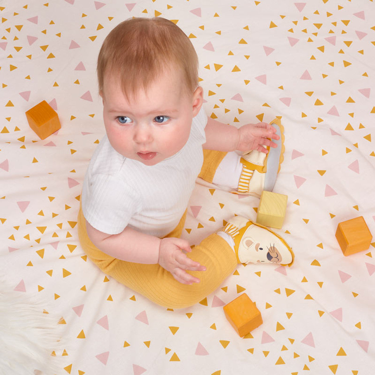 A unisex pram shoe by Start-Rite. Style is fable, a pull on in cream and yellow leather. Lifestyle picture of a baby sat on a mat wearing the pram shoes.