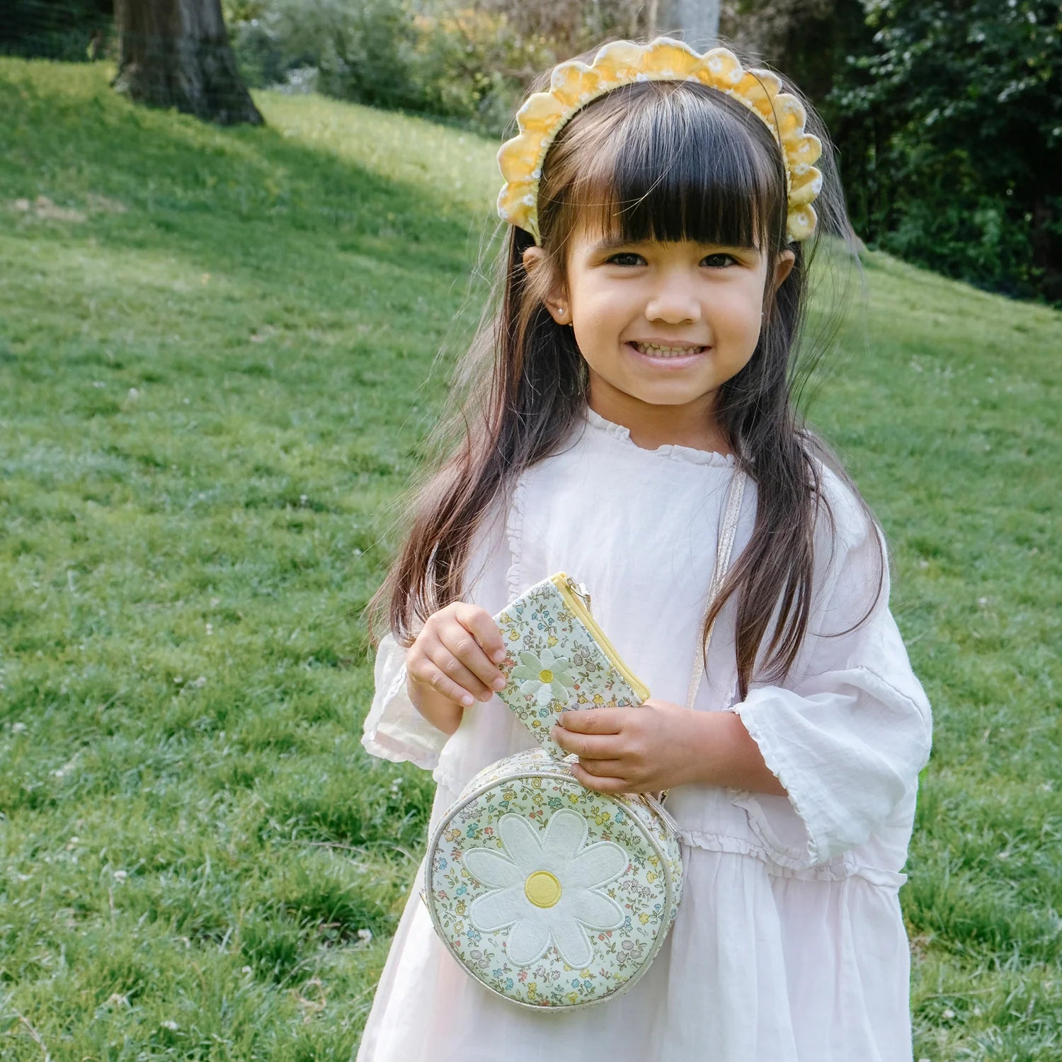 Young girl in a white dress with a floral headband holding a matching daisy bag by Rockahula outdoors.