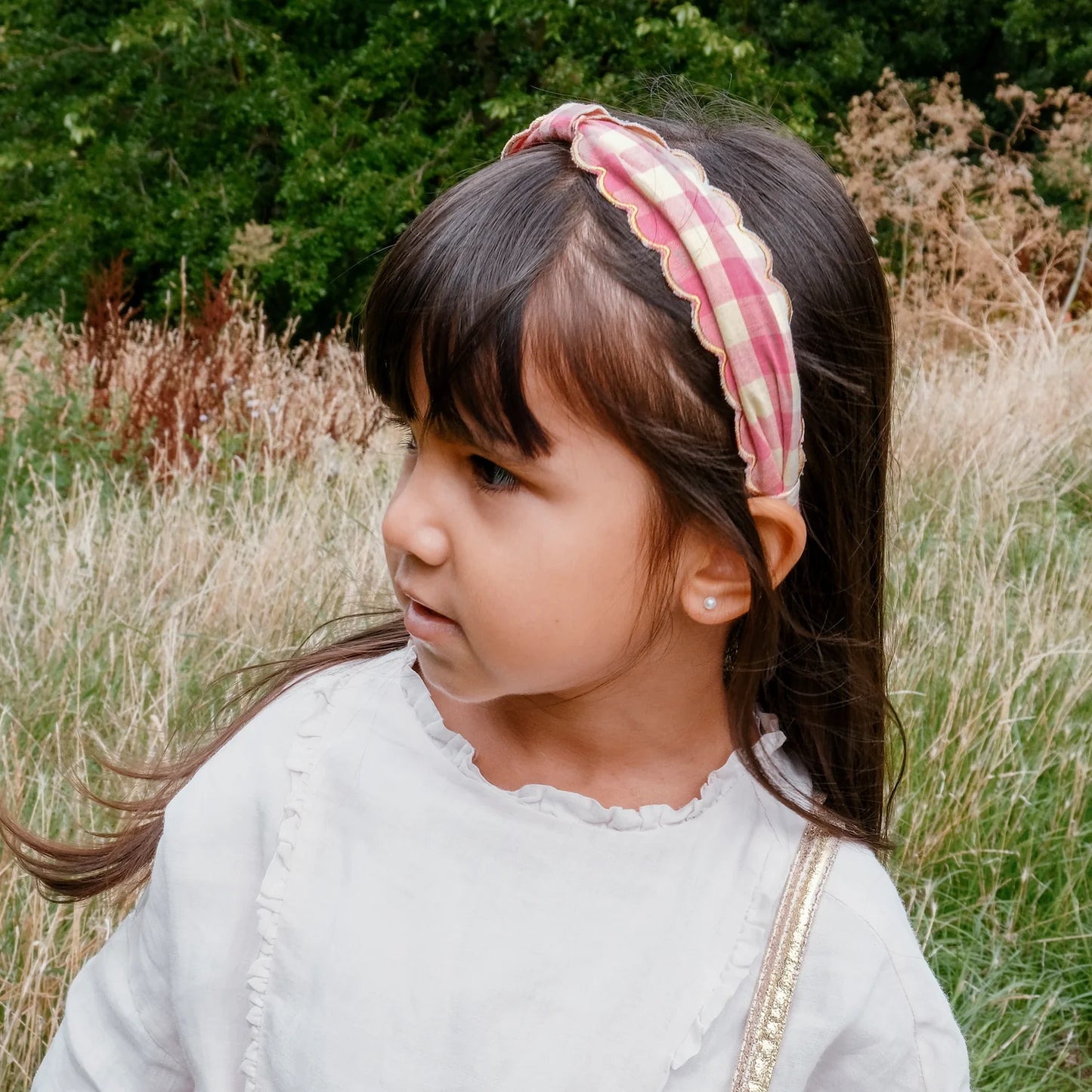 Young girl with a pink and white gingham headband by Rockahula standing in a natural setting with greenery and tall grass.
