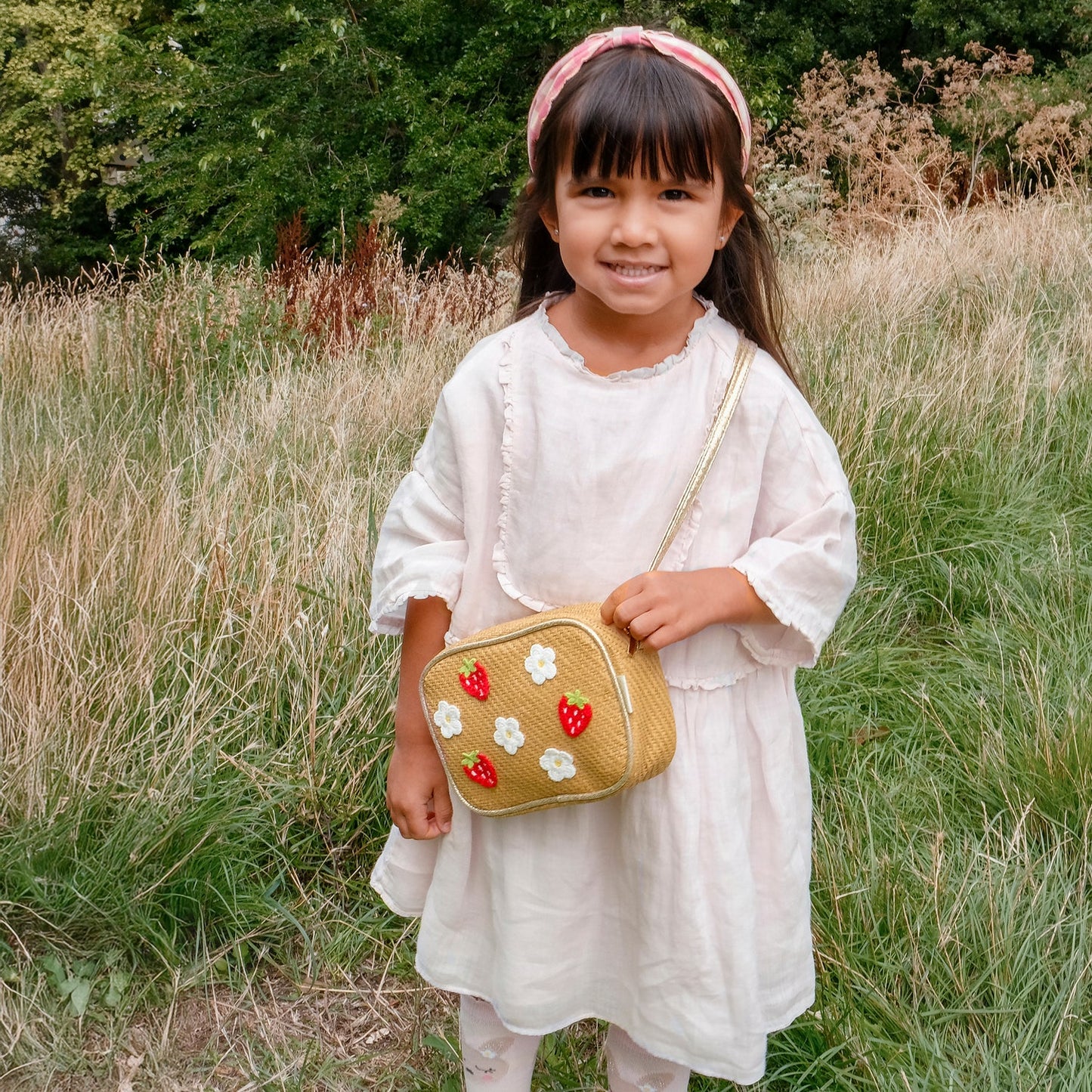 Young girl in a white dress and pink and white gingham headband by Rockahula, holding a gold purse with red and white flowers  in a grassy field.