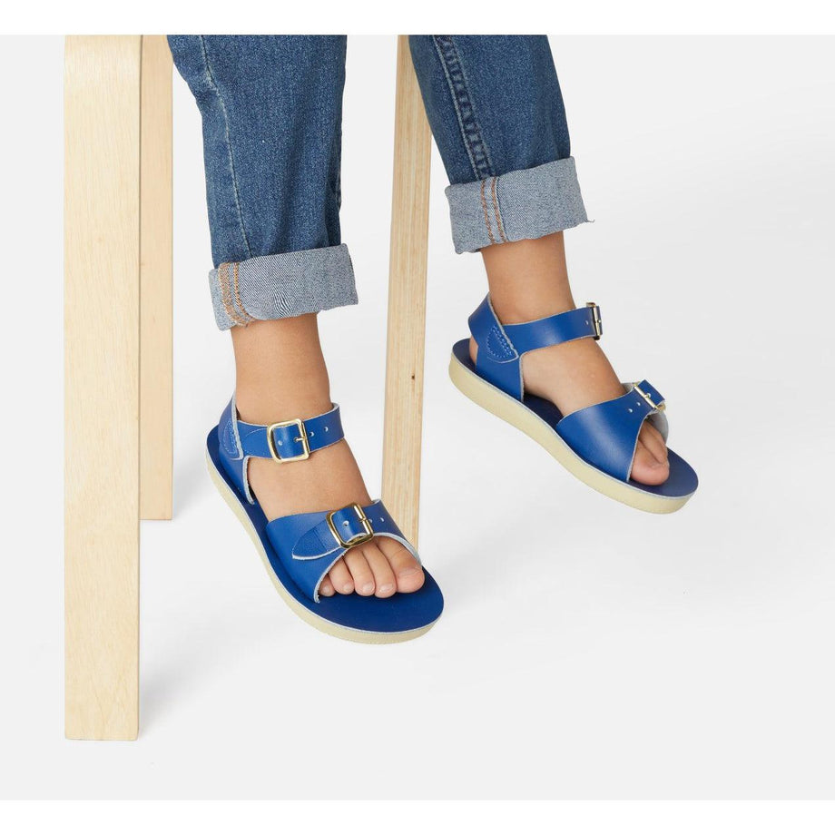 A unisex sandal by Salt Water Sandals in blue with double buckle fastening across the instep and around the ankle. Open Toe and Sling-back. Lifestyle of legs hanging off a chair view.