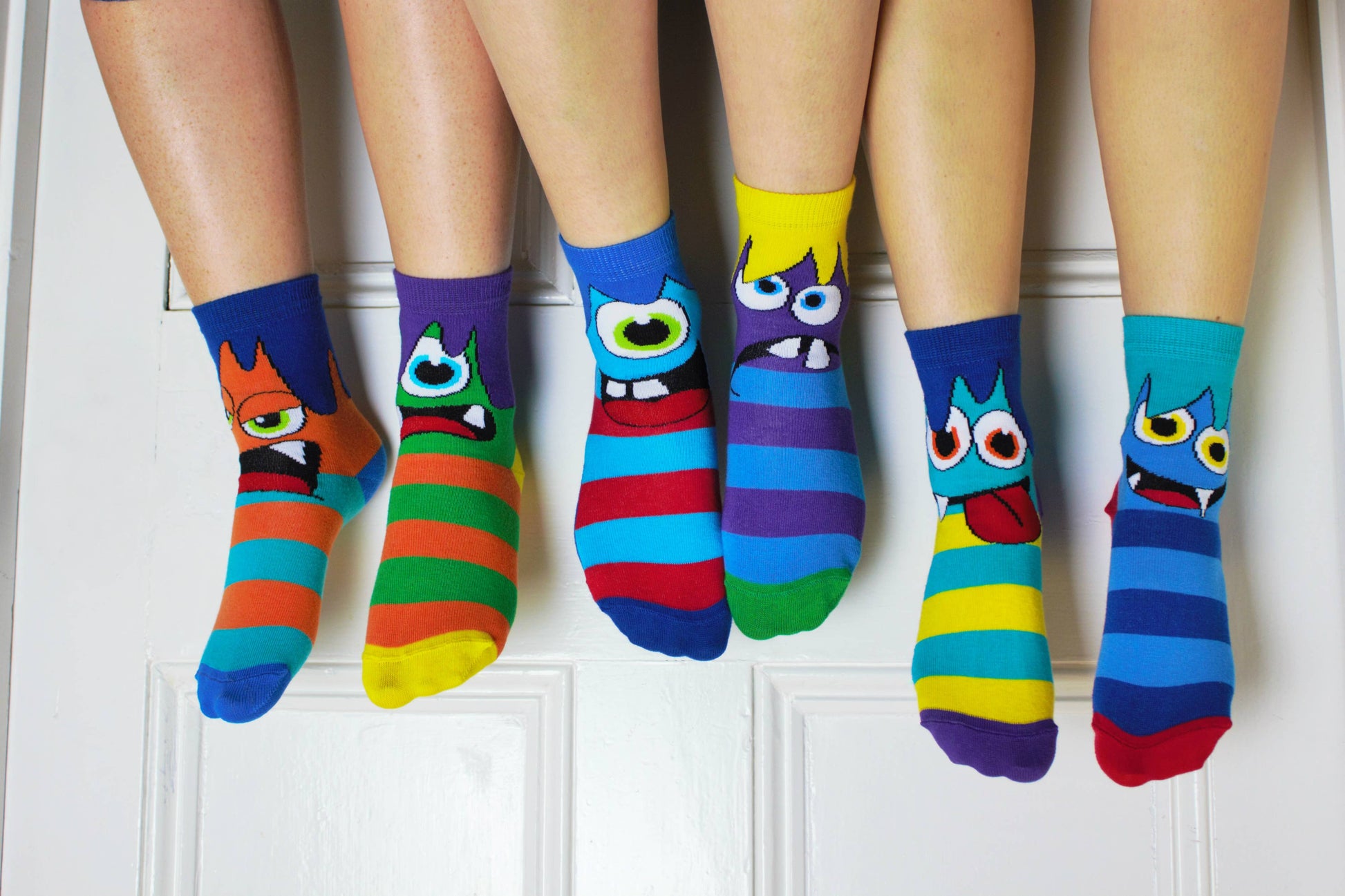 3 pairs of non matching multicoloured monster print socks by Odd Socks, style Mini Mashers. Front view of children wearing socks with a white door in the background.