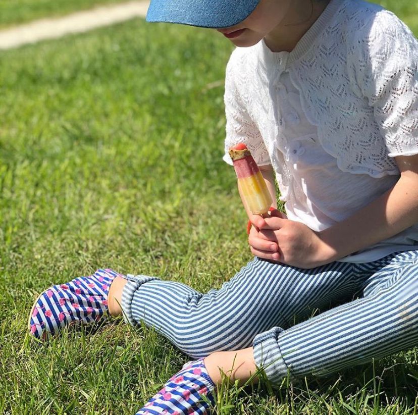 A girl sat on grass wearing a pair of non slip swim shoe by Slipfree, style Stripe, in blue, white, pink Flamingo. Angled view.