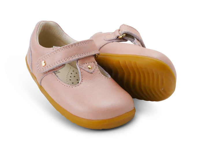 A girls first shoe by Bobux, style Louise, a velcro  T-bar in dusky pink pearl. Angled view of a pair, one showing the gum sole.