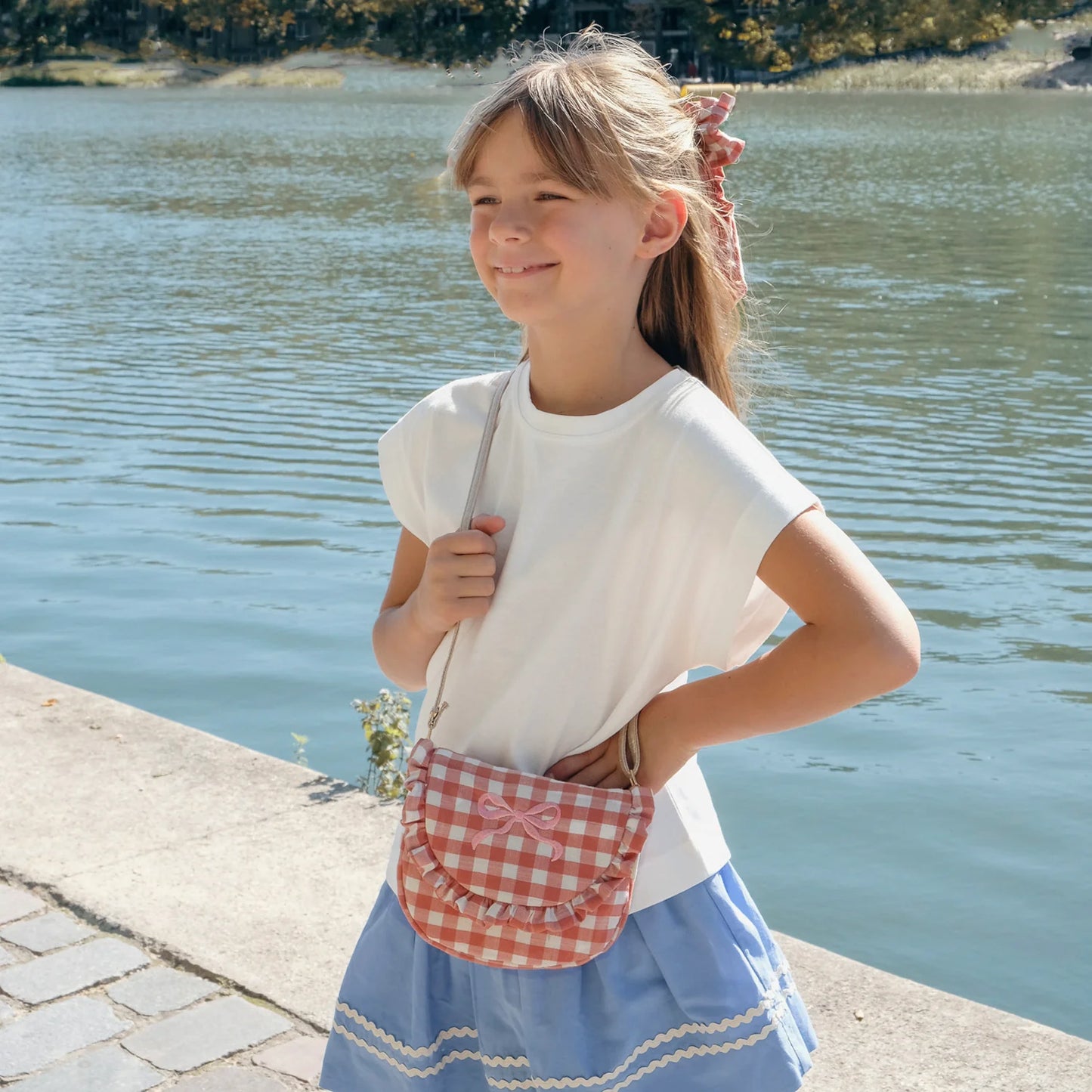 Young girl standing by a body of water wearing a white shirt and blue skirt with a pink and red gingham bag by Rockahula.