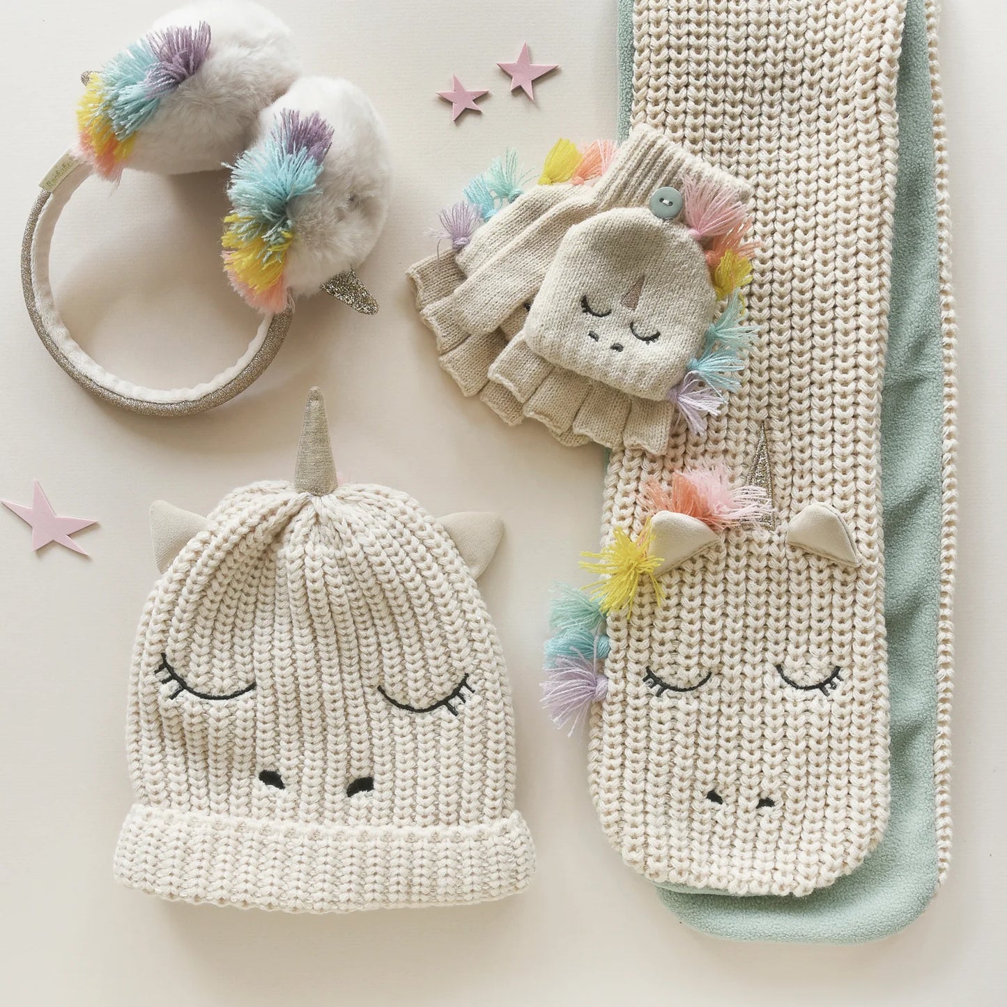 A pair of knitted fingerless gloves by Rockahula, style Rainbow Unicorn M2230M  in cream with fleece lined buttoned finger cap and embroidered unicorn face. Angled view showing matching accesories.