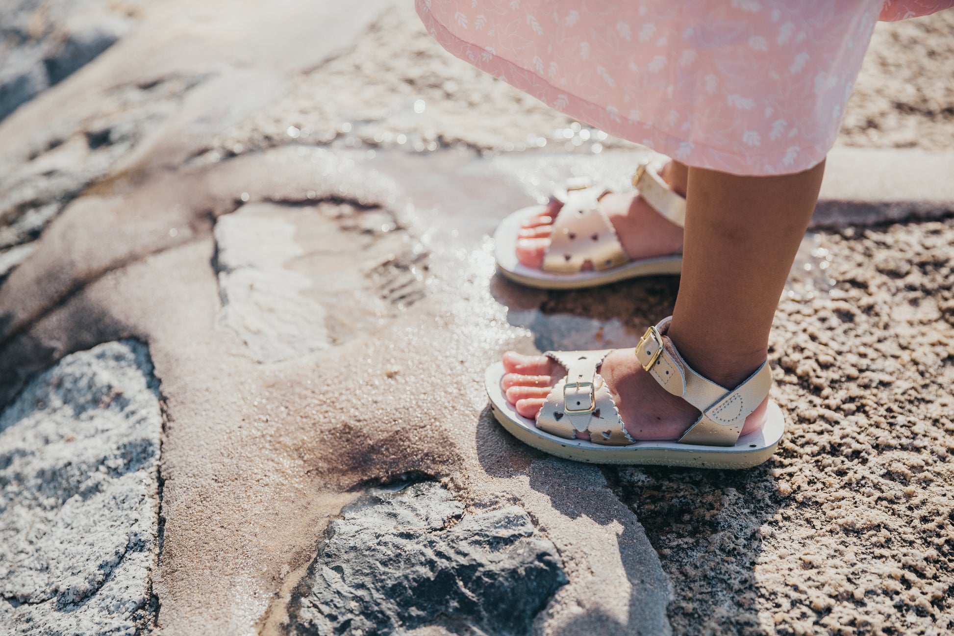 A girls sandal by Salt Water Sandal in rose gold with double buckle fastening across the instep and around the ankle. Featuring scallop edge and punched out heart detail. Lifestyle in water view.