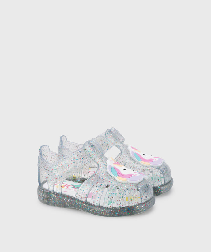 A girls jelly sandal by Igor, style Unicornio in transparent/multi/glitter with velcro strap and unicorn design on front. Angled side view of pair