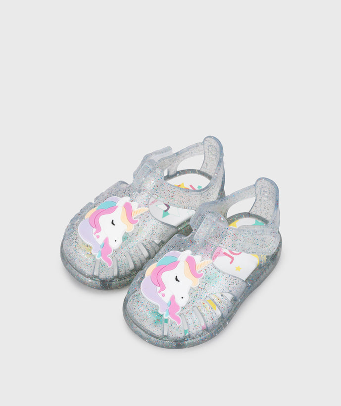 A girls jelly sandal by Igor, style Unicornio in transparent/multi/glitter with velcro strap and unicorn design on front. Angled front view of pair