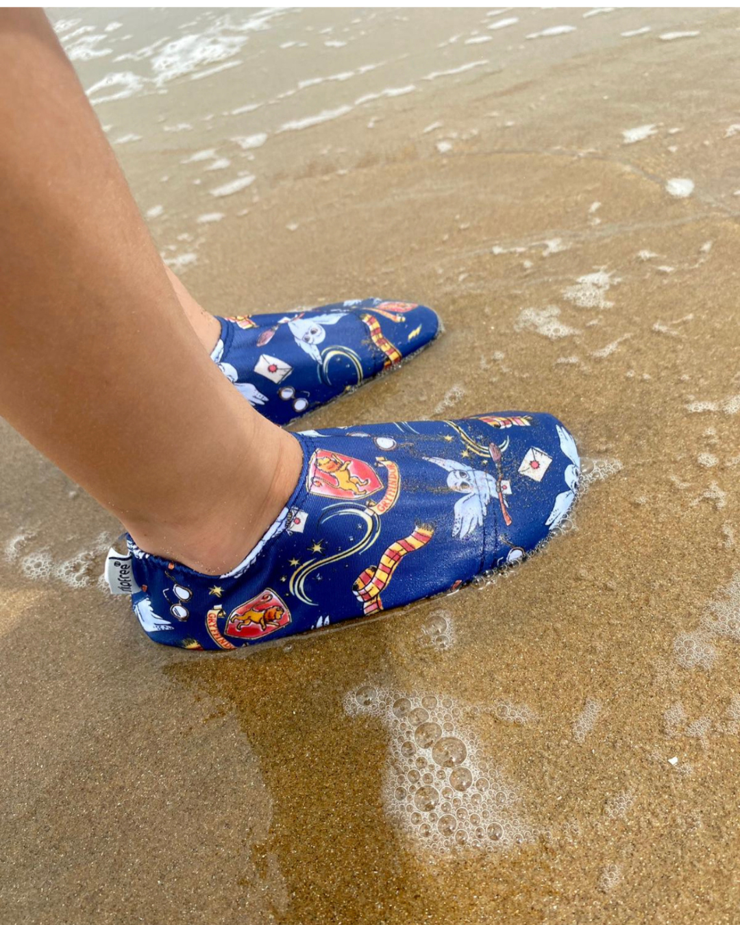 A unisex non slip shoe by Slipfree in colab with Warner Brothers, style Hedwig, in Harry Potter Owl print. Lifestyle image of feet in the sea.