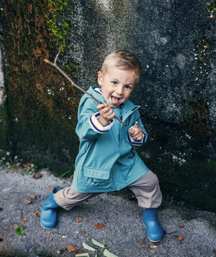 A hooded insulated raincoat by Igor, style Euri, with pockets and white and navy lining. Boy wearing raincoat playing with a stick.