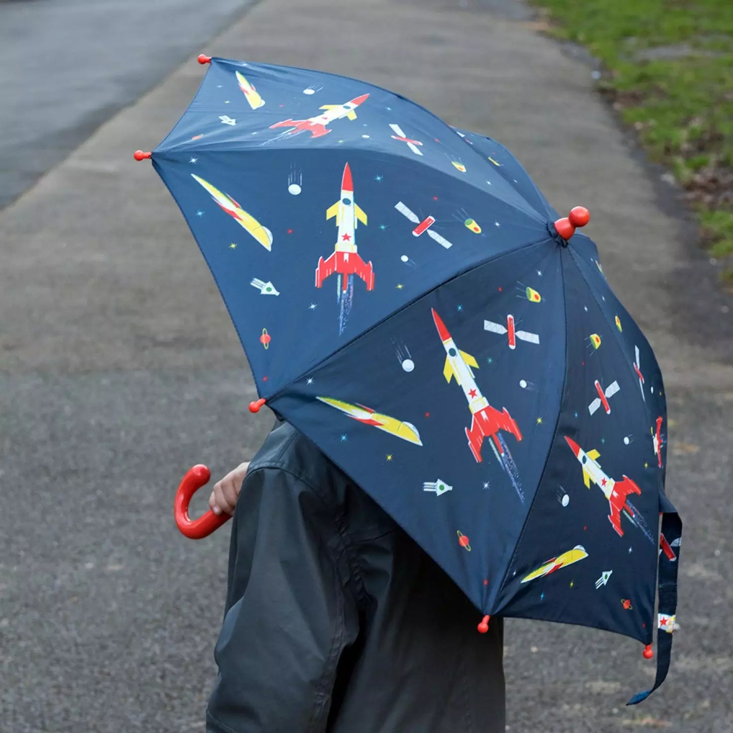 A childs umbrella by Rex London, style Space Age, in blue with multi rocket print and orange handle. Lifestyle image.