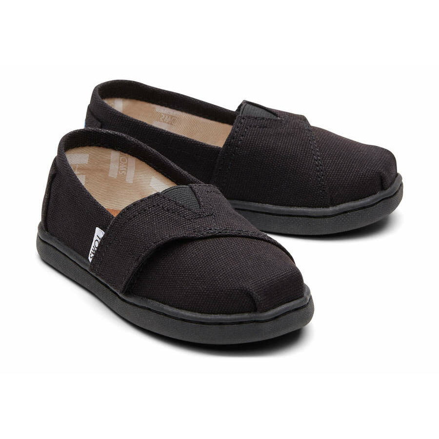 A pair of unisex canvas shoes by Toms, style Alpargata, in Black with velcro fastening. Angled view.