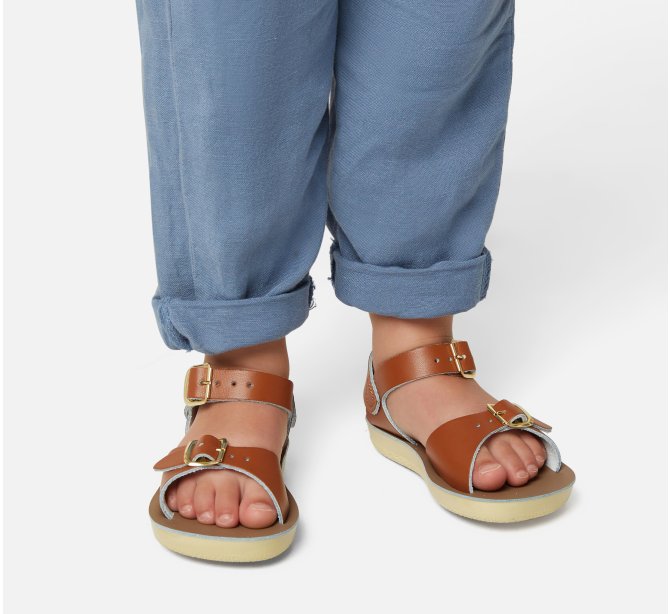 A unisex sandal by Salt Water Sandals in tan with double buckle fastening across the instep and around the ankle. Open Toe and Sling-back. Lifestyle of child's legs view.