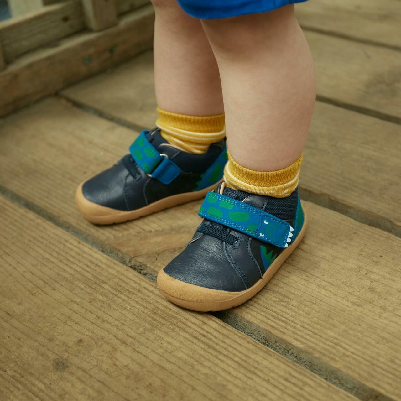 A wearing a pair of boys casual shoesw by Start Rite and JoJo Maman Bebé collaboration,style Companion,in Navy multi with dinosaur velcro fastening. Angled view.