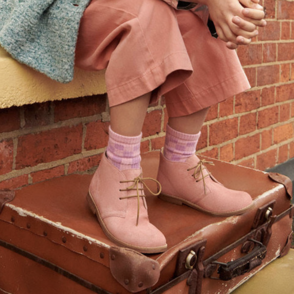 A girls desert boot by Start-Rite, style Autumn, in dusky pink glitter with light brown laces and soles. Girl wearing the boots resting on a suitcase.