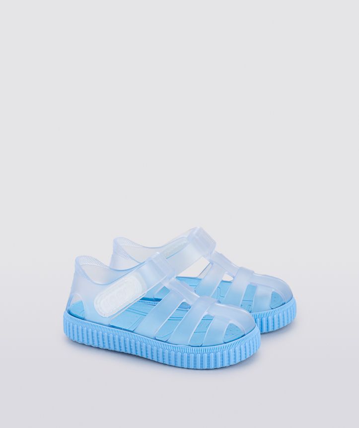 A unisex jelly shoe by Igor, style Nico Cristal, in clear with a blue sole and velcro strap. Angled view.